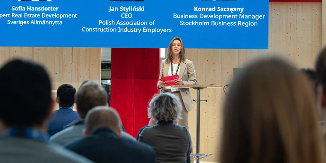  kvinnlig moderator står på scen och talar till en engagerad publik under en livepresentation på Nordbygg.